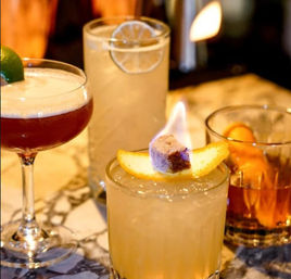 Assorted craft cocktails on a marble bar top: frothy ruby coupe with lime, tall citrus highball with a lime wheel, crushed-ice rocks glass topped with a flaming sugar cube and orange slice, and an amber old-fashioned with orange peel.