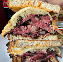 Close-up of a stacked New York-style deli pastrami sandwich: thick smoked pastrami slices, sauerkraut, melted Swiss cheese and grilled rye bread with charred edges and juices visible.