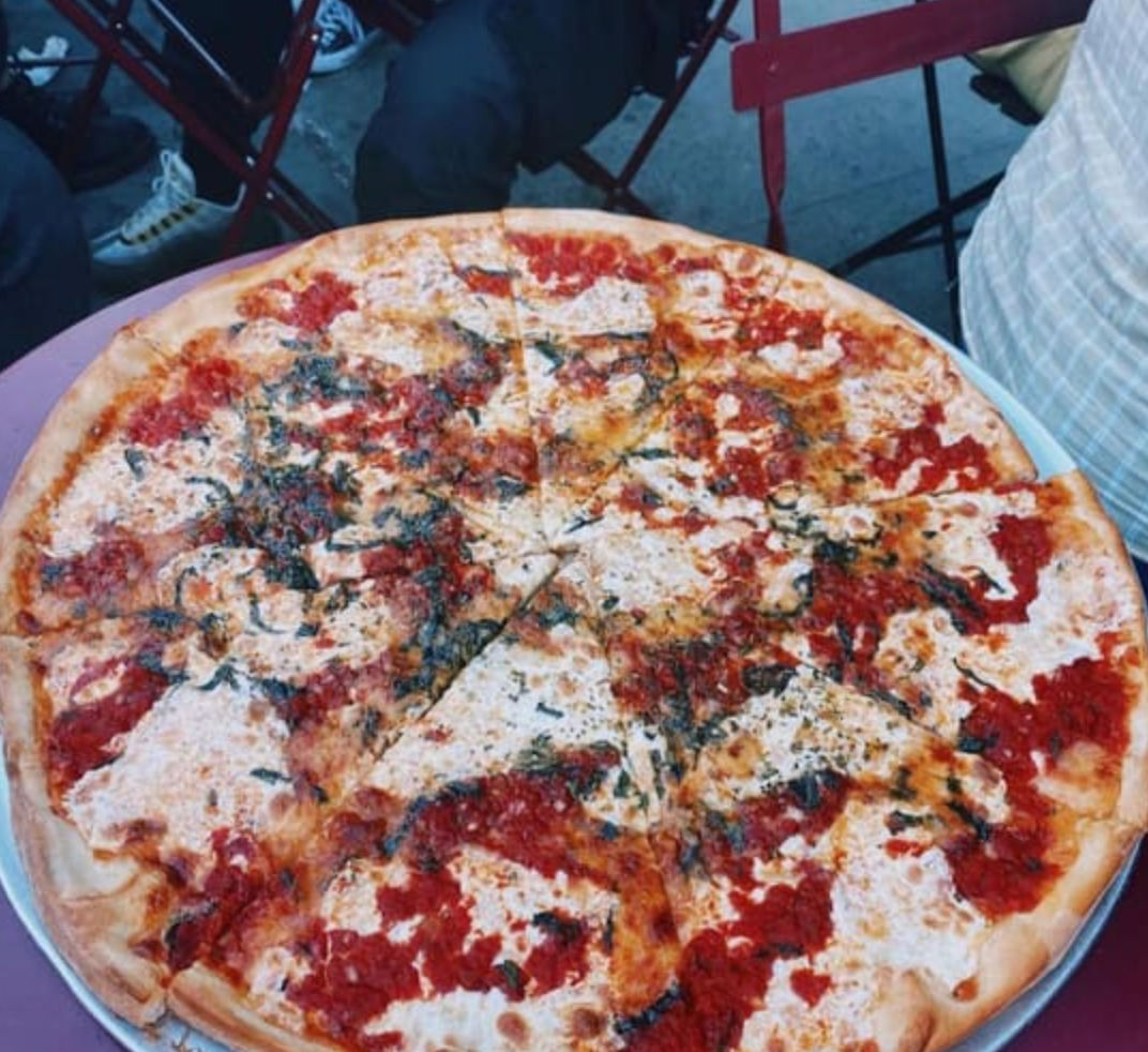 Large thin-crust margherita-style pizza with melted mozzarella, dollops of tomato sauce and basil on a metal tray at an outdoor café table