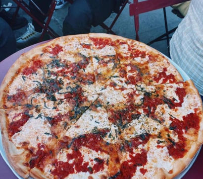 Large thin-crust margherita-style pizza with melted mozzarella, dollops of tomato sauce and basil on a metal tray at an outdoor café table