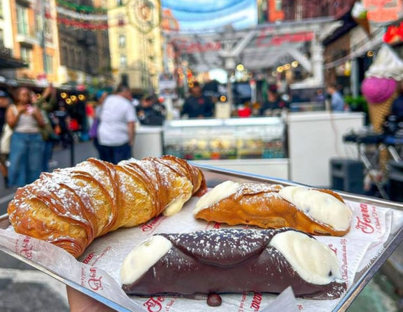 Handheld tray of pastries at a bustling outdoor street market: a powdered-sugar croissant and two cream-filled cannoli (one chocolate-dipped) with blurred shoppers and market stalls in the background.