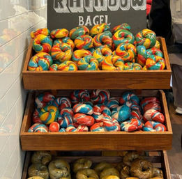 Bright rainbow swirl bagels piled on wooden bakery display trays, vivid blue, red and yellow marbled bagels on upper shelves with green-tinted bagels below
