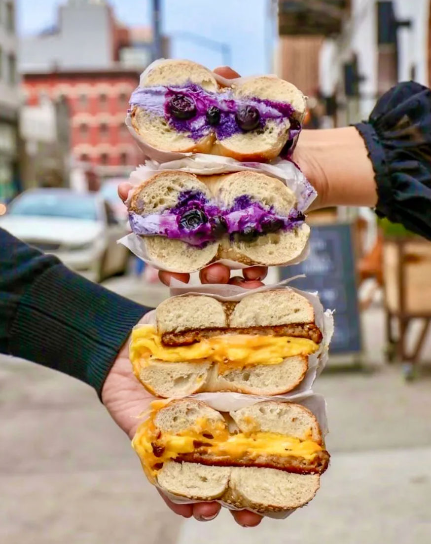 Hand-held stack of four bagel sandwiches on a city street: two blueberry cream-cheese bagels with fresh berries and vivid purple spread, and two gooey melted-cheese breakfast bagels with egg and crispy patty.