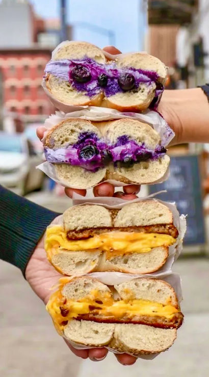 Hand-held stack of four bagel sandwiches on a city street: two blueberry cream-cheese bagels with fresh berries and vivid purple spread, and two gooey melted-cheese breakfast bagels with egg and crispy patty.