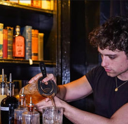 Bartender pouring a brown craft cocktail through a strainer into an ice-filled glass at a dimly lit bar, with bottles and books on a shelf in the background.