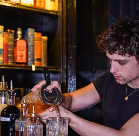 Bartender pouring a brown craft cocktail through a strainer into an ice-filled glass at a dimly lit bar, with bottles and books on a shelf in the background.