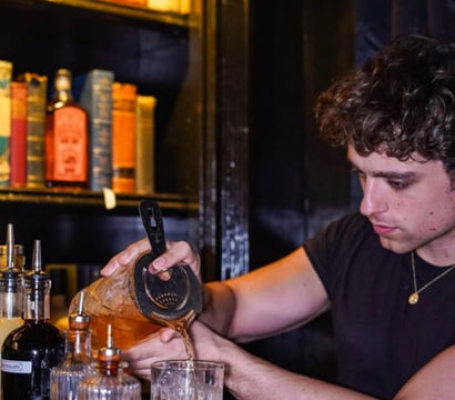 Bartender pouring a brown craft cocktail through a strainer into an ice-filled glass at a dimly lit bar, with bottles and books on a shelf in the background.