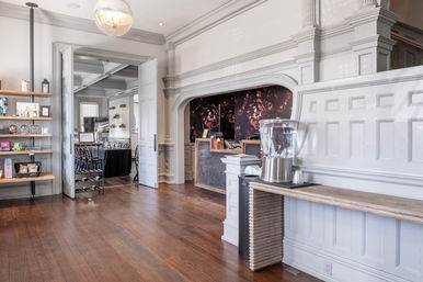 Sunlit historic hotel reception with ornate white paneling and dark wood floors, open double doors to a dining area, a shelving display of snacks and a self‑serve water dispenser on a rustic counter.