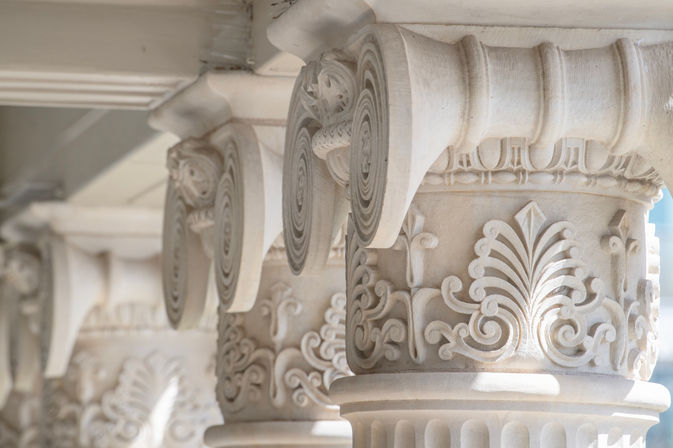 Close-up of sunlit classical stone column capitals with carved volutes and acanthus leaf motifs in a row, ornate architectural detail on a historic building facade