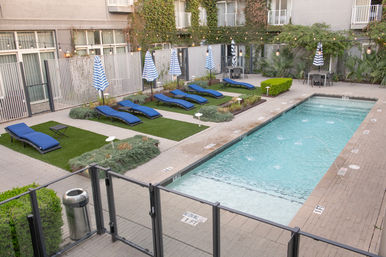 Modern urban courtyard outdoor pool with rectangular lap pool, blue lounge chairs, striped umbrellas, string lights and ivy-covered building balconies.