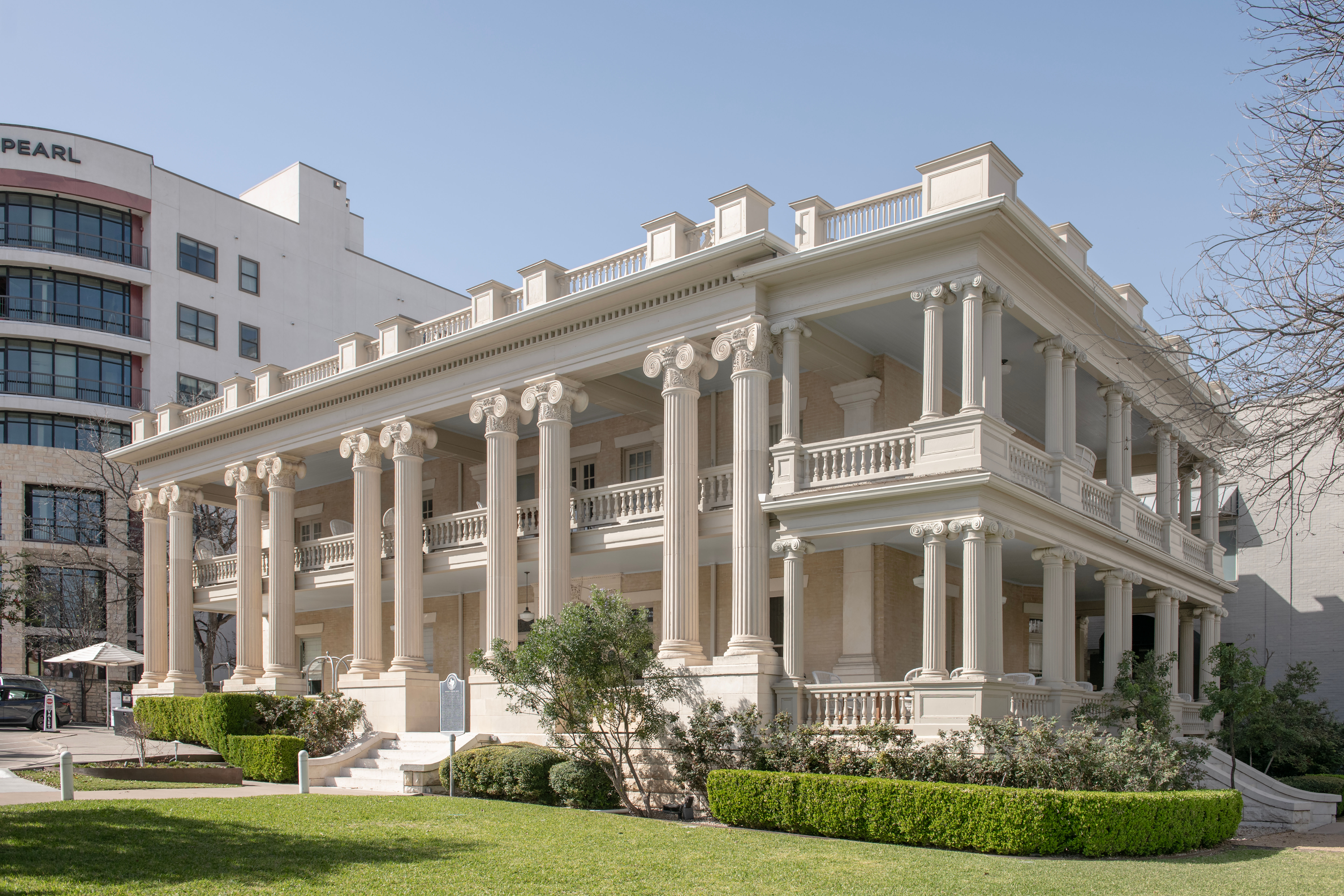 Sunlit historic neoclassical mansion with two-story colonnade of Corinthian columns, wraparound porches and balustrades, manicured hedges and lawn in an urban setting