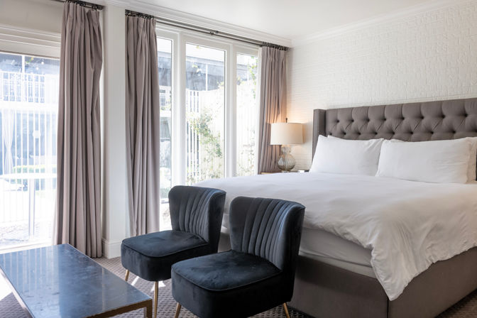 Bright modern hotel-style bedroom with king bed and tufted grey headboard, crisp white linens, two navy velvet chairs and floor-to-ceiling glass doors to a sunny courtyard