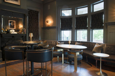 Cozy upscale lounge interior with curved tufted banquette, round marble tables, black leather bar chairs, brass-accent lamps, bay windows with shades, and hardwood floors.