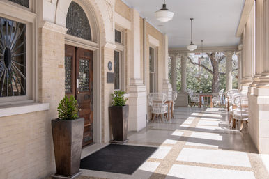 Sunlit historic porch with arched wooden door, classical columns, potted plants and wicker chairs arranged for outdoor dining