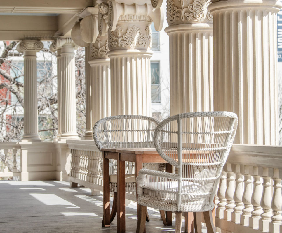Sunlit porch with ornate classical columns, two white wicker chairs and a small wooden table on a balustraded veranda.