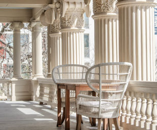 Sunlit porch with ornate classical columns, two white wicker chairs and a small wooden table on a balustraded veranda.
