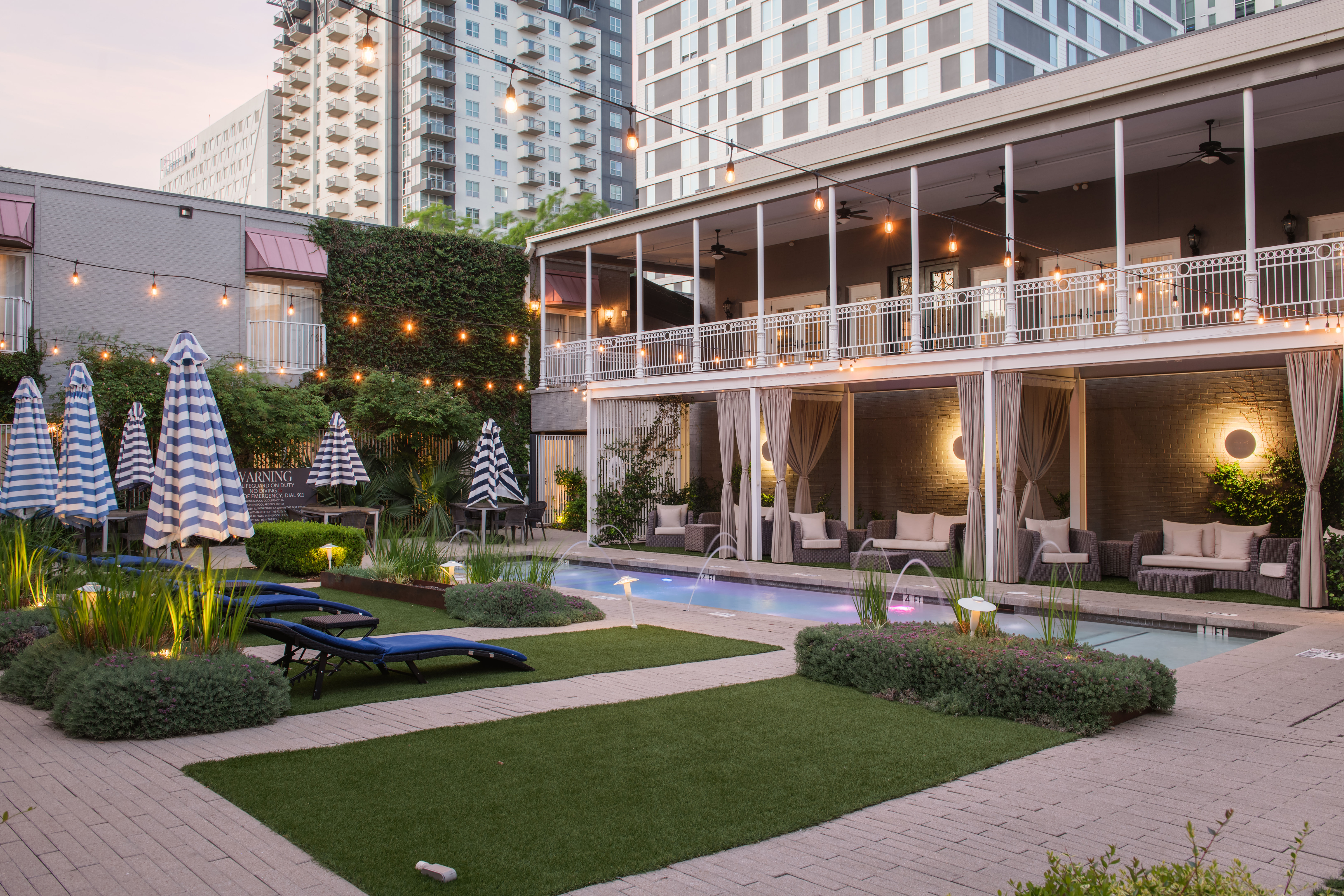 Urban hotel courtyard at dusk with a narrow lap pool, curtained cabanas, striped umbrellas, blue lounge chairs, string lights and high-rise buildings