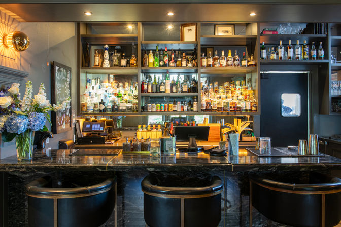 Upscale cocktail bar interior with black marble countertop, three leather bar stools, backlit shelves full of liquor bottles, cocktail tools and a vase of blue and white flowers.
