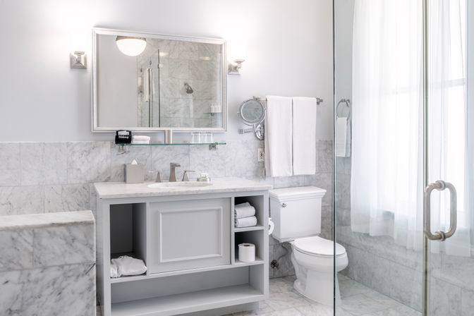 Bright spa-like white marble bathroom with marble floor and walls, glass shower door, gray vanity with open shelves, chrome fixtures, toilet, and neatly folded towels by a sunlit window