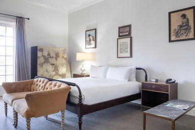 Sunlit boutique bedroom with dark wooden spindle bed dressed in crisp white linens, tufted tan loveseat at the foot, nightstands with lamp and phone, framed artwork on pale walls, marble-top coffee table and large curtained window.