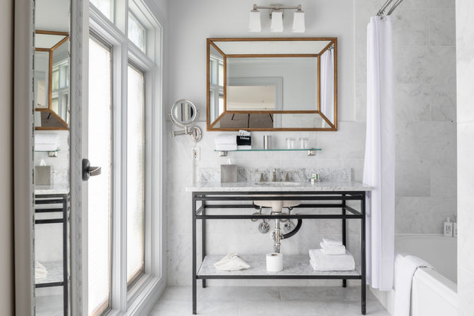 Sunlit spa-style white marble bathroom with black metal vanity and marble countertop, gold-framed mirror, wall magnifier, glass shelf, folded towels and tub with white shower curtain.