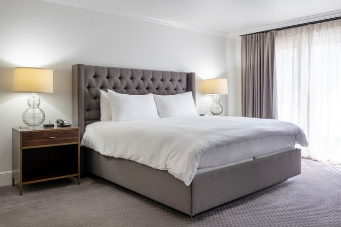 Bright, inviting hotel bedroom with king-size tufted gray headboard, crisp white bedding, glass bedside lamps on wooden nightstands, and floor-to-ceiling curtains filtering soft natural light.