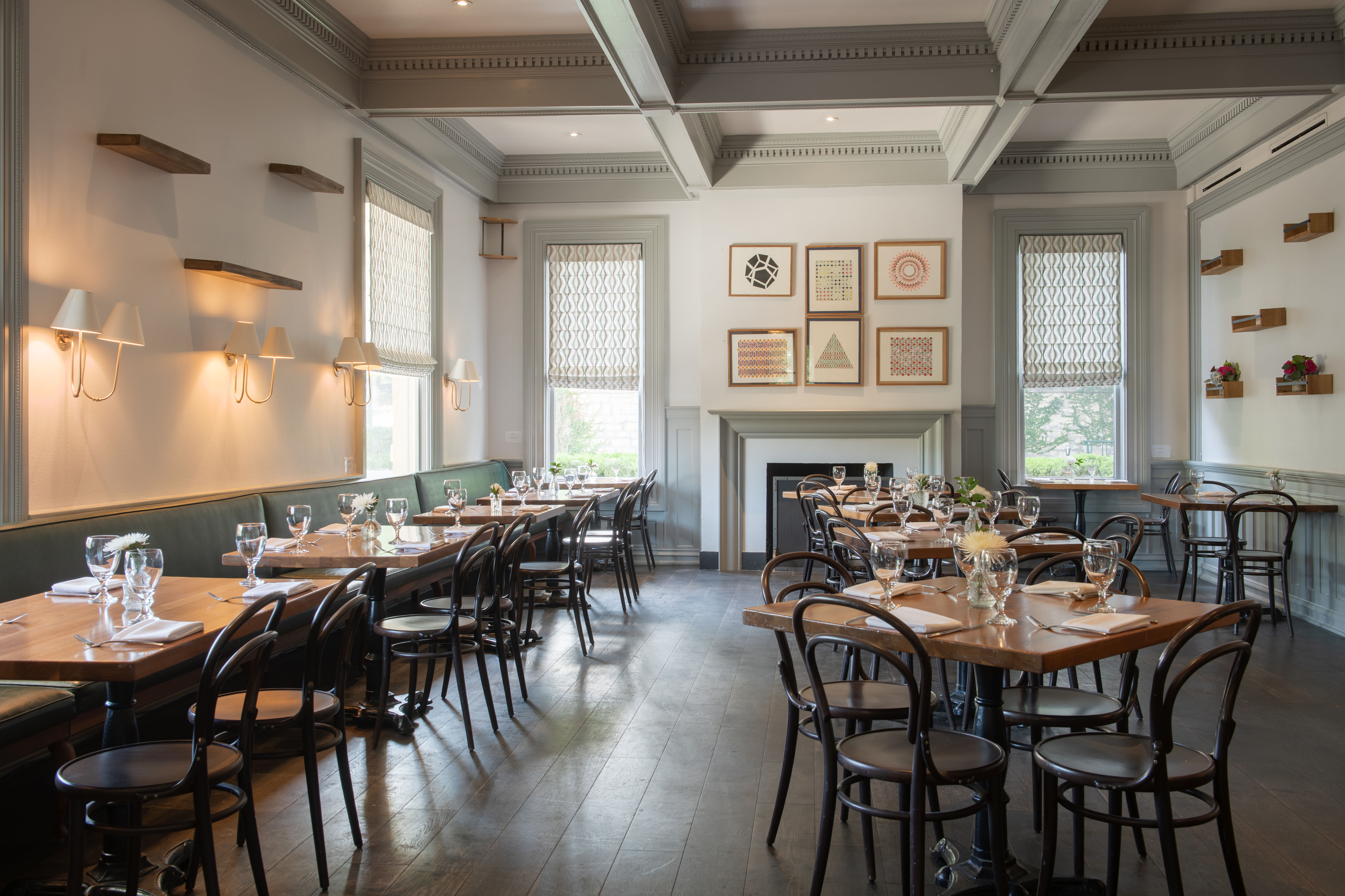 Bright, airy bistro dining room with wooden tables and bentwood chairs, green banquette seating, patterned Roman shades, wall sconces and framed geometric art; tables set with glassware and small floral centerpieces, inviting and contemporary