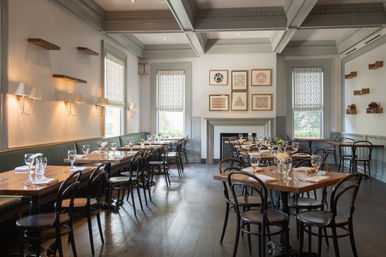 Bright, airy bistro dining room with wooden tables and bentwood chairs, green banquette seating, patterned Roman shades, wall sconces and framed geometric art; tables set with glassware and small floral centerpieces, inviting and contemporary