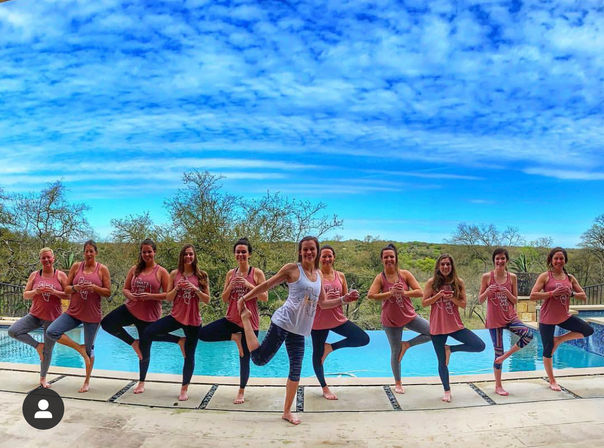 Poolside group yoga: a line of women in matching pink tanks (one in white) practicing tree pose on an outdoor pool deck with bright blue sky and wooded hillside beyond.