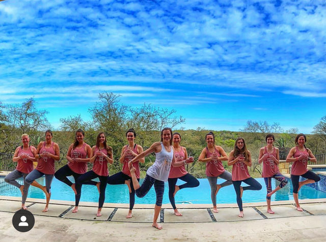 Poolside group yoga: a line of women in matching pink tanks (one in white) practicing tree pose on an outdoor pool deck with bright blue sky and wooded hillside beyond.