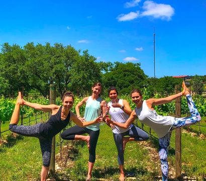 Vineyard yoga group of four women practicing tree and dancer poses while clinking wine glasses in a sunny grapevine row under a bright blue sky