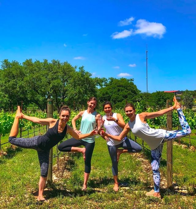 Vineyard yoga group of four women practicing tree and dancer poses while clinking wine glasses in a sunny grapevine row under a bright blue sky