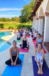 Smiling group yoga class on colorful mats poolside at a sunny Mediterranean-style patio, with curved swimming pool, green lawn and trees in the background.