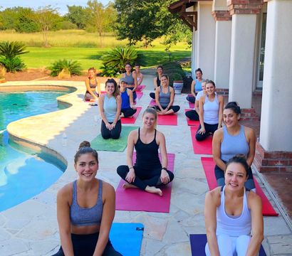 Smiling group yoga class on colorful mats poolside at a sunny Mediterranean-style patio, with curved swimming pool, green lawn and trees in the background.