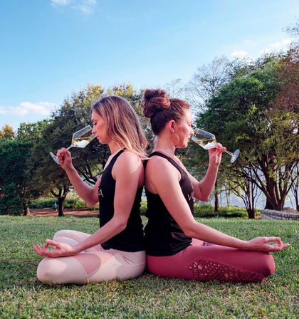 Two women seated back-to-back on grass in a sunny park, wearing yoga leggings and tank tops and holding wine glasses while meditating — playful outdoor yoga and wine scene.