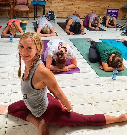 Smiling participant in pigeon pose at an airy indoor yoga studio, surrounded by a group practicing child's pose on mats — water bottles and a wooden wall create a cozy class vibe.