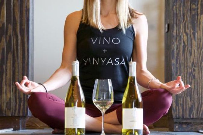 Person seated cross-legged in an indoor yoga space wearing a tank top reading VINO + VINYASA, with two white wine bottles and a wine glass in front — playful wine-and-yoga lifestyle image.