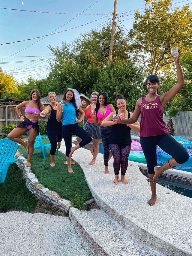 Seven women in colorful activewear doing yoga tree pose and holding wine glasses at a backyard pool bachelorette-style party with inflatables and patio chairs at sunset.