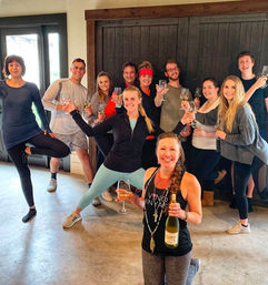 Energetic group of adults in activewear posing and toasting with wine glasses in a rustic tasting room, front woman kneeling and holding a bottle of white wine.