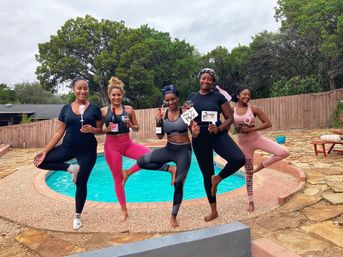 Five smiling friends in activewear doing yoga tree poses by a backyard swimming pool, holding champagne bottles and bachelorette signs for a playful outdoor celebration.