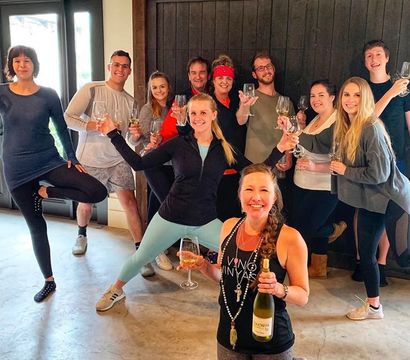 Lively group wine tasting: about a dozen friends in casual wear cheering with wine glasses inside a cozy winery tasting room with rustic wood doors, woman in front holding a bottle.