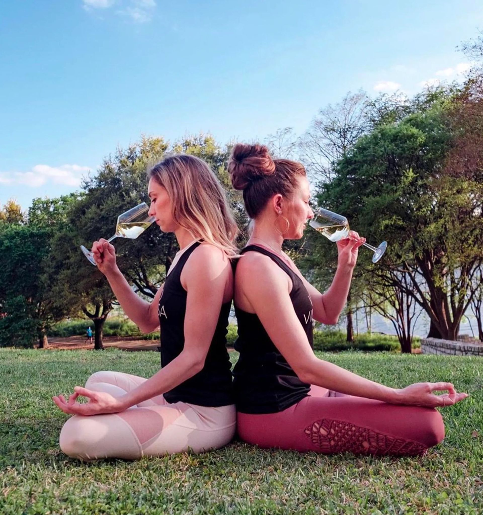 Two women sit back-to-back in a cross‑legged yoga pose on a grassy park lawn, each holding a glass of white wine to their nose, wearing athletic leggings and tank tops with trees and blue sky behind them.