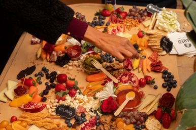Hand reaching for a colorful charcuterie grazing board with assorted cheeses, cured meats, strawberries, grapes, pomegranate, nuts, crackers, mini peppers and a honey dip — festive party snack spread.