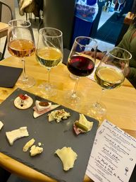 Four-glass wine flight (rosé, two whites, red) with a slate cheese and charcuterie board on a wooden table at a cozy indoor wine bar, menu visible
