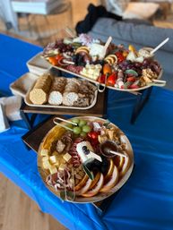 Colorful charcuterie spread on wooden platters with cheeses, cured meats, crackers, grapes, berries, citrus slices and honey on a blue tablecloth — inviting party grazing board.