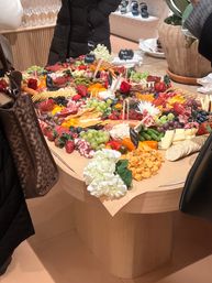 Gourmet charcuterie grazing table on a round wooden display with cheeses, cured meats, grapes, strawberries, crackers, pickles, nuts, dips and small chalkboard labels, decorated with white hydrangea and shoppers browsing nearby