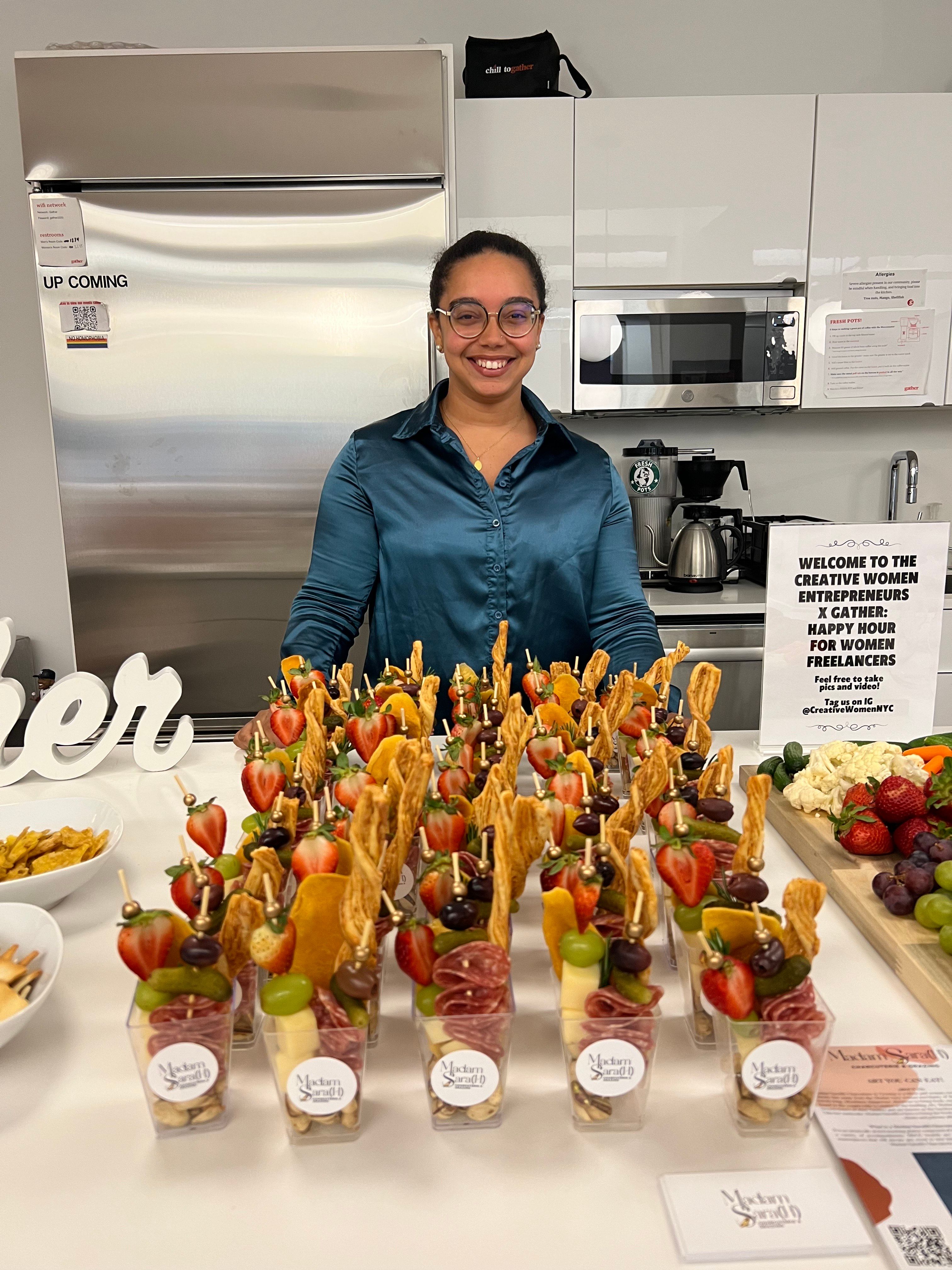 Smiling woman behind a display of individual charcuterie cups with strawberries, grapes, cheese, cured meats and breadsticks at a women’s entrepreneurs networking event in a modern kitchenette.