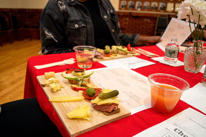 Wooden tasting board with assorted cheeses, cured meats, olives, cornichon and sliced fruit on a red tablecloth, plastic cup of juice and menus at an indoor tasting event.