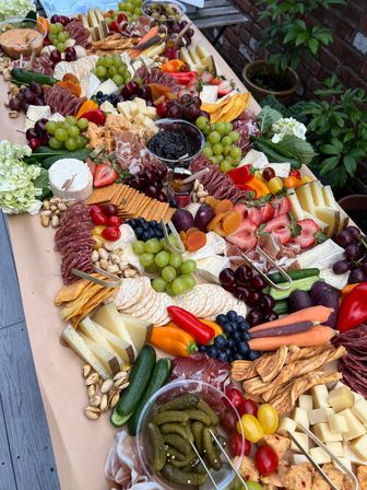 Vibrant outdoor charcuterie grazing board on a paper-lined table in a garden — assorted cheeses, cured meats, crackers, grapes, strawberries, dried apricots, olives, cornichons, nuts, mini peppers, cucumbers and dips.