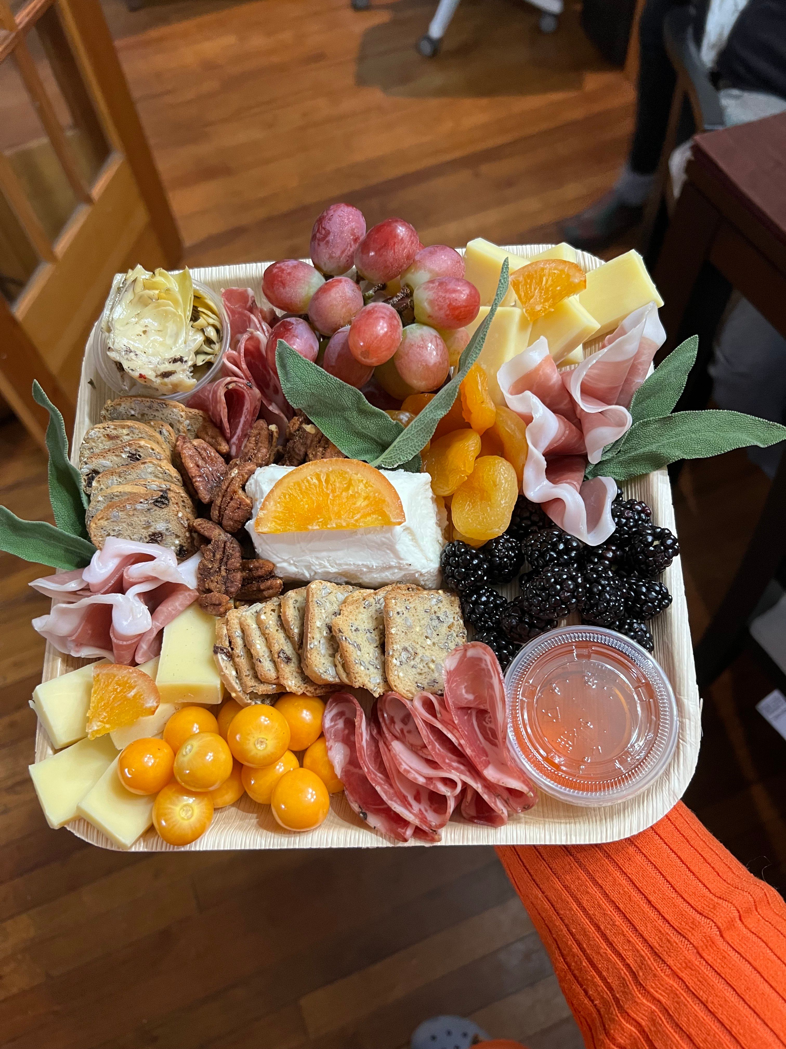 Artisan charcuterie grazing box with brie topped by an orange slice, cubed cheese, salami and prosciutto, crackers, grapes, blackberries, dried apricots and a honey dip — handheld in a cozy home kitchen.